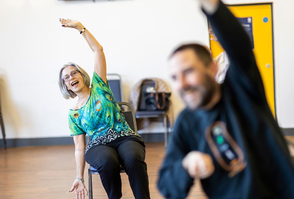 An elderly woman is sat down performing a stretch with her arms over her head.