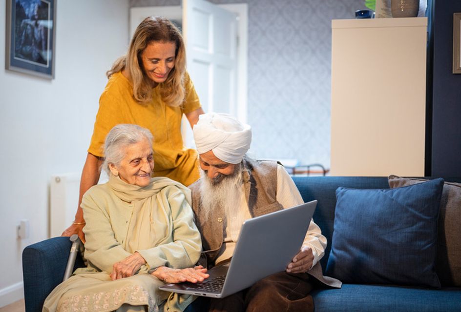 An older Asian couple are sat on a sofa looking at a laptop. A younger woman stands behind them, also looking at the laptop.
