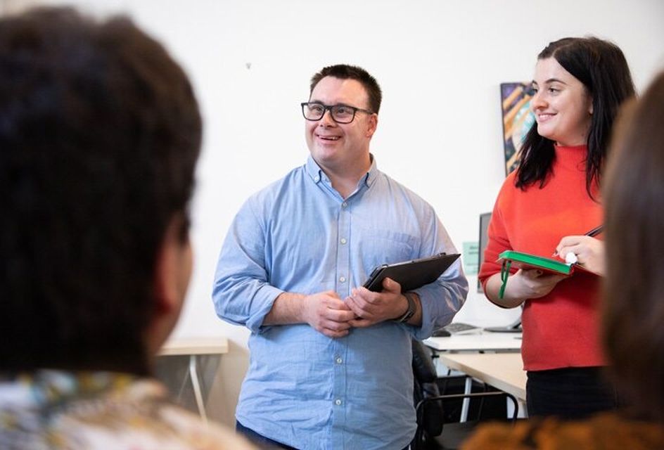A man holding a clipboard smiles at his colleagues as they talk in an office setting. The man is white and wears blue shirt and glasses. Next to him is a woman with brown hair and a red top.