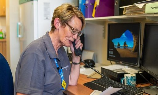 NHS staff member on the phone at a desk, reading a document in an office setting.