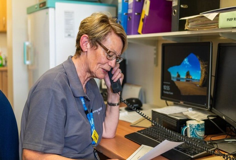 NHS staff member on the phone at a desk, reading a document in an office setting.