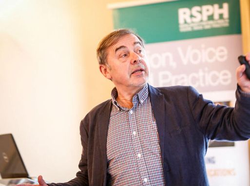 A white teacher with grey hair speaks at the front of a classroom