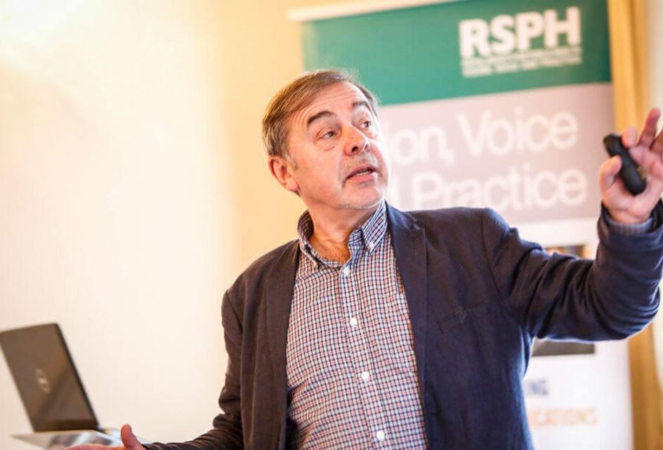 A white teacher with grey hair speaks at the front of a classroom