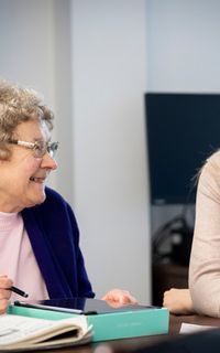 An elderly white woman with glasses writes on a notepad as she talks to a care worker