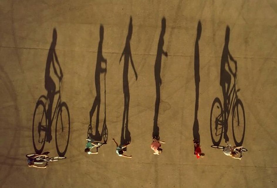 Shadows of people cycling, scooting and roller skating stretch out across a playing field.