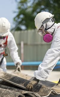 Two people wearing protective suits removing a roof panel containing asbestos.