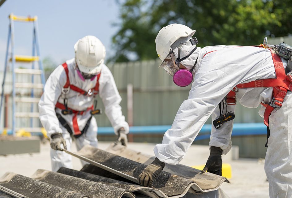 Two people wearing protective suits removing a roof panel containing asbestos.