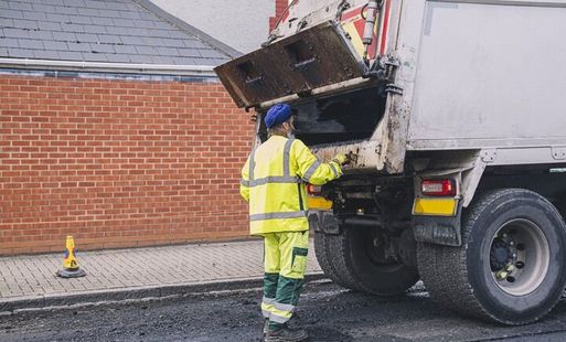 A refuse collector collects rubbish and puts it into a van. He is wearing high vis clothing.
