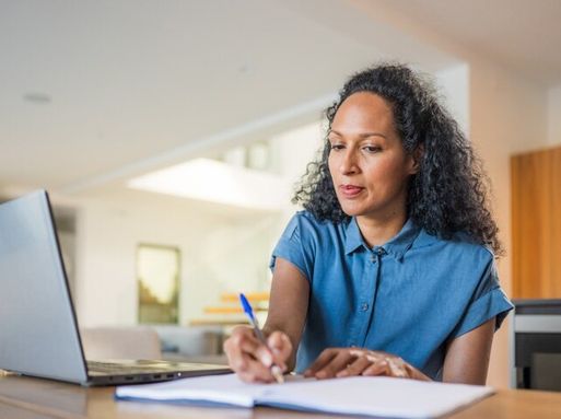 A woman is writing on a notepad with a laptop next to her.