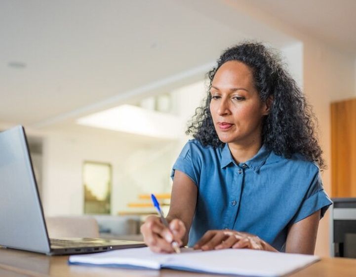 A woman is writing on a notepad with a laptop next to her.