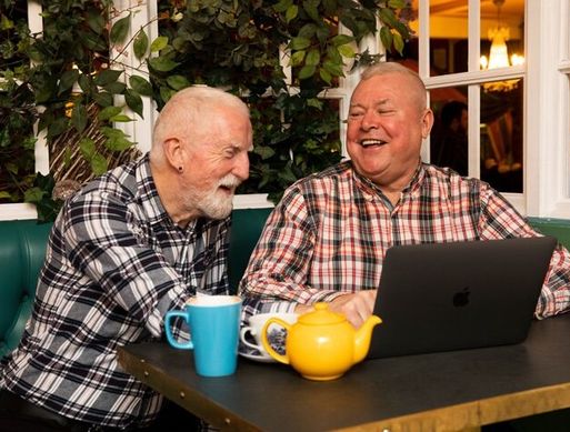 Two elderly white men sit at a laptop with a pot of tea laughing and talking