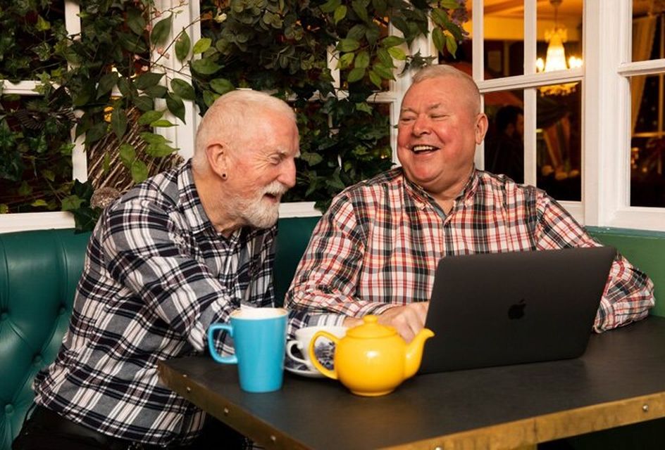 Two elderly white men sit at a laptop with a pot of tea laughing and talking