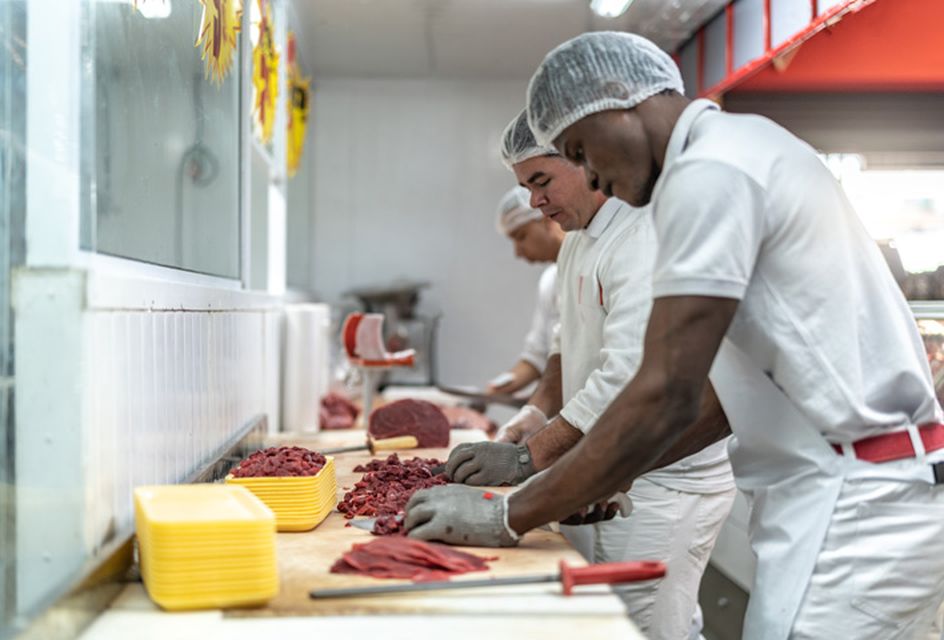 A line of chefs wearing aprons and hair nets chopping meat in a kitchen.
