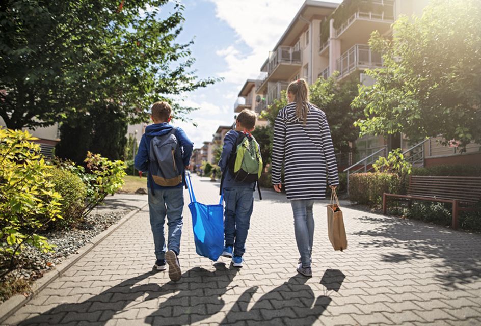Two young boys carry a plastic bag between them. To their right, their mother walks alongside them.