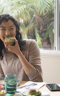 An Asian woman with black curly hair smiles while eating a piece of toast with avocado on it.
