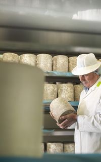 A cheesemonger wearing a white coat and a white hat inspects a wheel of cheese in a factory.