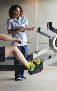 A physio assists a man as he uses a rowing machine.