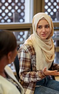 A woman wearing a hijab and plaid shirt speaks to a group of people.