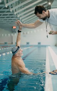 An elderly man in a swimming pool high-fives a swimming instructor standing on the side of the pool
