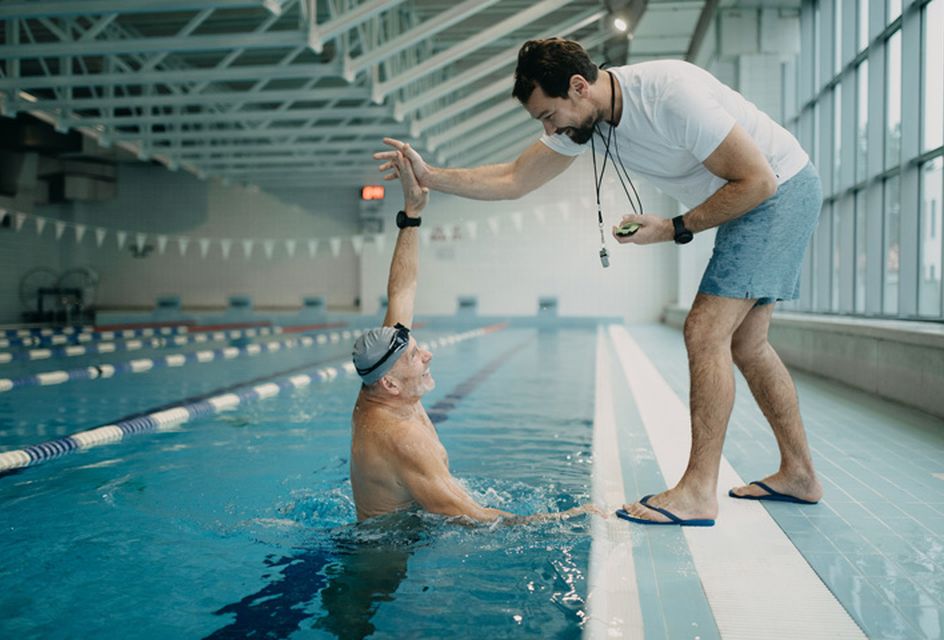 An elderly man in a swimming pool high-fives a swimming instructor standing on the side of the pool