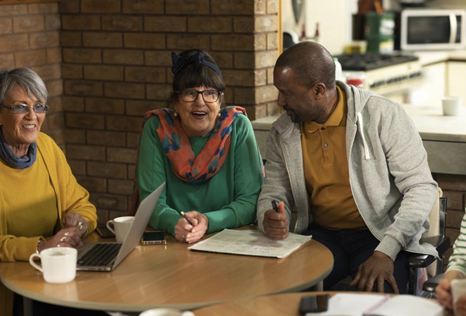 Two women and a man sit at a round table, warmly smiling and engaged in conversation. A laptop and cups are on the table