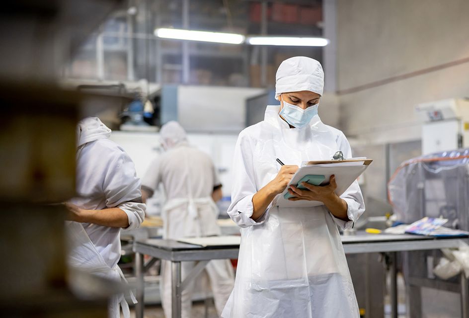 A woman in a white apron, hat and face mask takes notes on a clipboard in a facotyr.