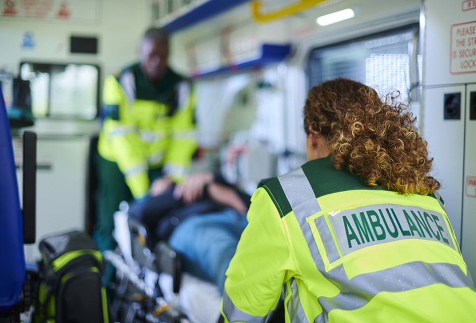 A paramedic wearing a high-vis that reads "ambulance" pushes a patient in a stretcher into an ambulance.