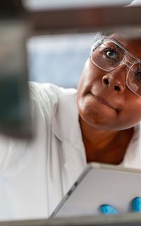 A woman in a white coat, hair net and goggles inspects a piece of machinery in a factory and takes notes on a clipboard.