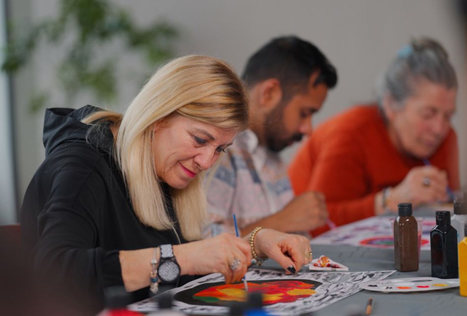 A white woman with blonde hair paints a colourful picture. Two others are in the background painting too.