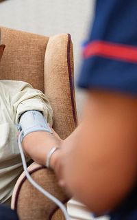 An older man gets his blood pressure checked by a nurse. He is sat in an armchair.