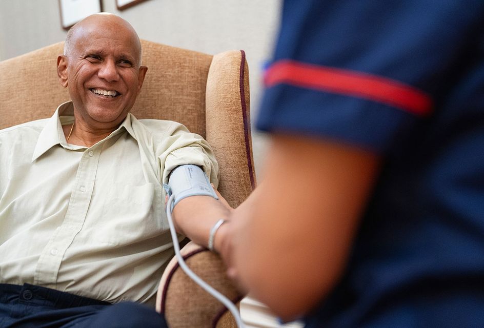 An older man gets his blood pressure checked by a nurse. He is sat in an armchair.
