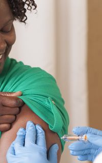 A black woman with short curly hair smiles while getting a vaccine.