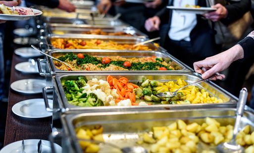 People serve themselves food from a buffet with trays of vegetables, potatoes, and other dishes. Plates and serving utensils are visible.