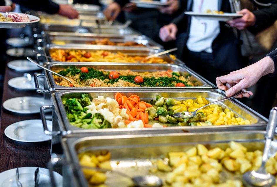 People serve themselves food from a buffet with trays of vegetables, potatoes, and other dishes. Plates and serving utensils are visible.
