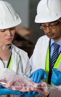 Two technicians in white coats, high vis jackets and hard hats inspect a vacuum-sealed bag of meat.