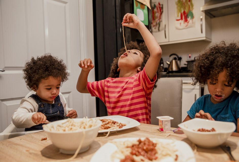 Three young children eating spaghetti at a kitchen table.