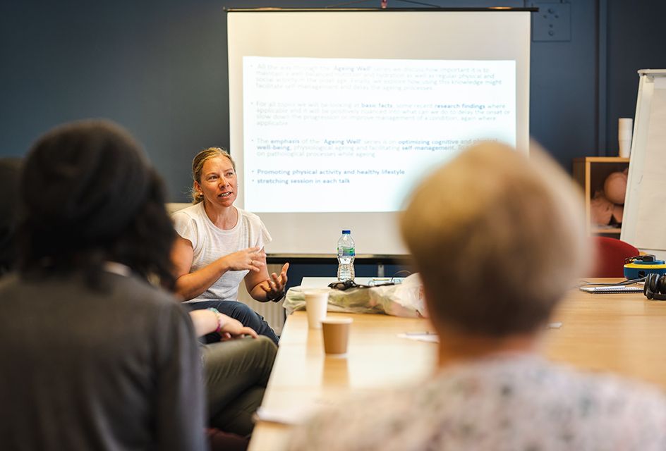 A trainer sits at the front of a classroom speaking in front of a presentation.