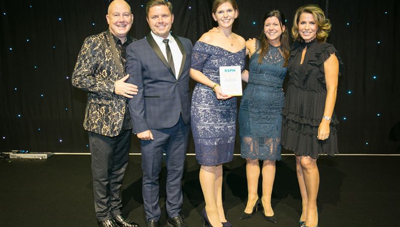 Five people stood on stage with three middle accepting an Award from Natasha Kaplinsky