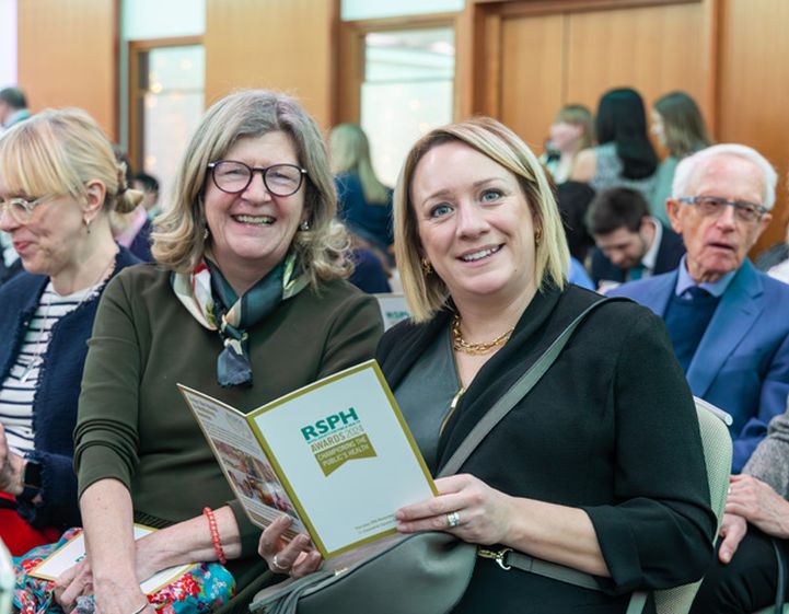 Two women sitting down at a busy Health & Wellbeing  Awards ceremony smiling to camera with one holding an event programme.