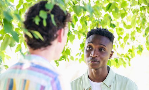 A man looking sympathetically at another man under a leafy tree on a sunny day.