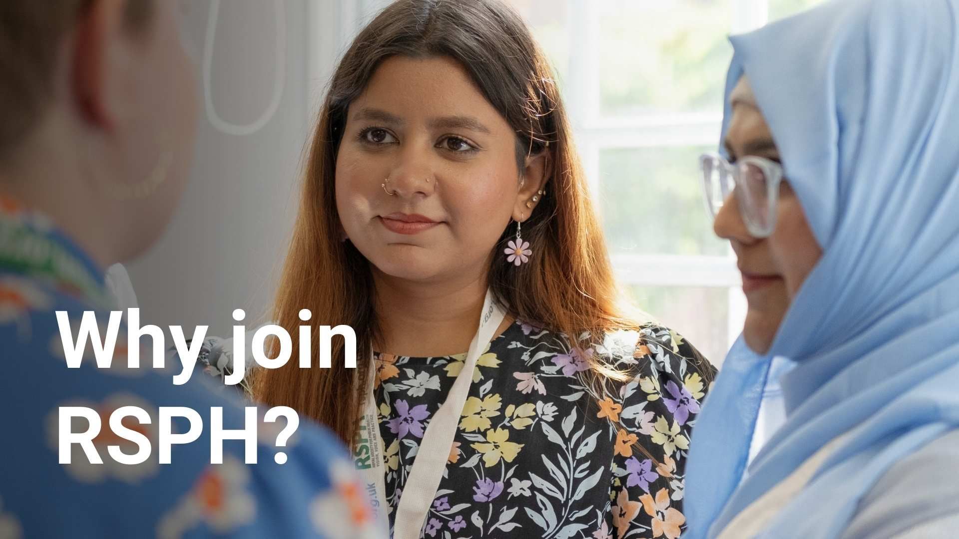 Two young women listen to another woman who is talking in the foreground. They are both wearing RSPH lanyards.