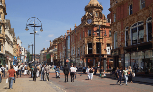 A bustling Leeds city centre street with pedestrians, lined with historic buildings featuring ornate architecture and a clock tower