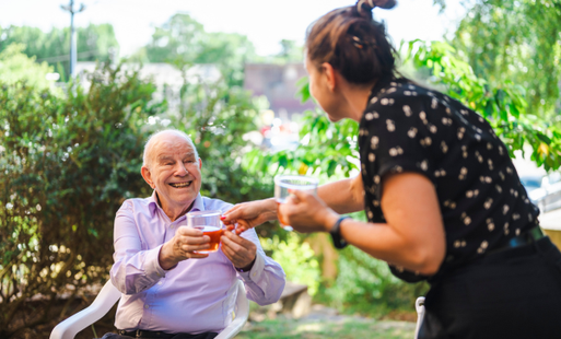 Smiling elderly man receiving a drink from a woman in a sunny garden setting.